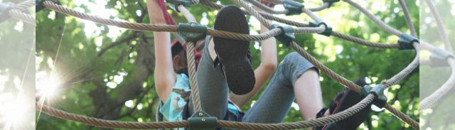 Ein kleines Mädchen klettert ein Kletternetz hoch. Sie trägt einen Helm, ein Top und ¾-Leggings mit Sandalen.