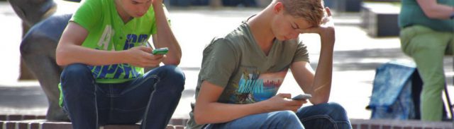 Chiropraktik im Einsatz gegen den Handynacken Zwei Jugendliche sitzen auf einer Steinmauer und gucken in ihr Smartphone. In der einen Hand halten sie ihr Smartphone, mit der anderen stützen sie ihren Kopf.