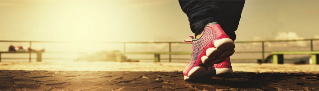 Nahaufnahme von Turnschuhe an Füßen, die in Richtung einer Promenade gehen. Der Untergrund ist mit Steinplatten versehen.