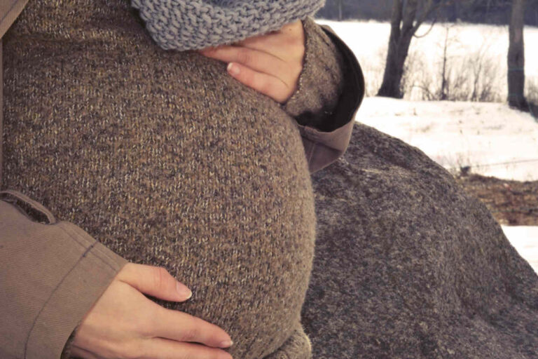 Nahaufnahme eines Schwangerschaftsbauches. Die Frau trägt einen braunen Pullover und einen braunen Mantel.