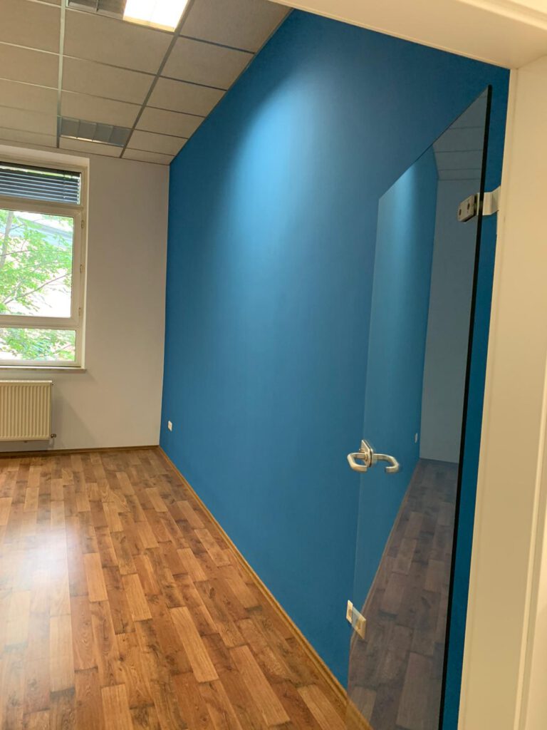 Eingang eines Raumes mit einer blauen Wand, rechts des Einganges. Die Eingangstür ist aus Glas.