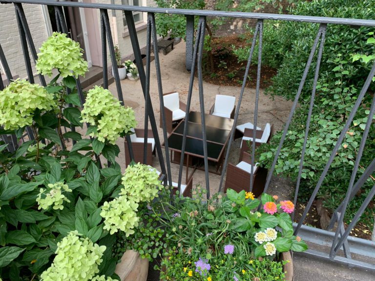 Auf einem Balkon mit Gitterstäben stehen mehrere Blumentöpfe. Unten in einem Garten steht eine braune Sitzgarnitur mit weißen Polstern.