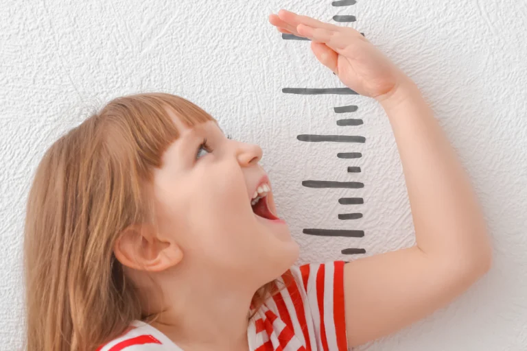 Ein kleines Mädchen hält ihre Hand an ein selbtgemaltes Maßband, um ihre Größe zu messen. Dabei lacht sie glücklich