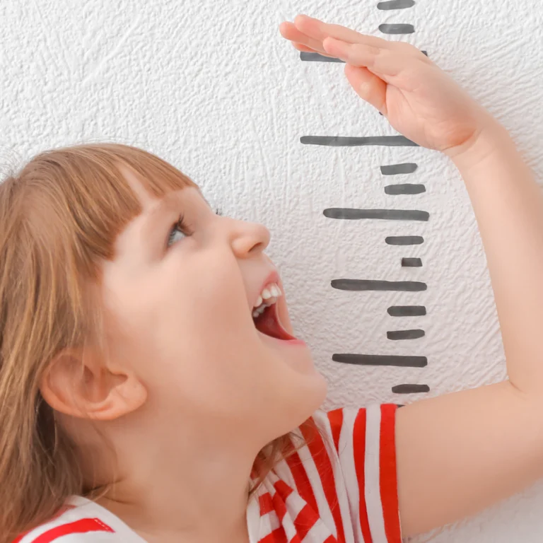 Ein kleines Mädchen hält ihre Hand an ein selbtgemaltes Maßband, um ihre Größe zu messen. Dabei lacht sie glücklich