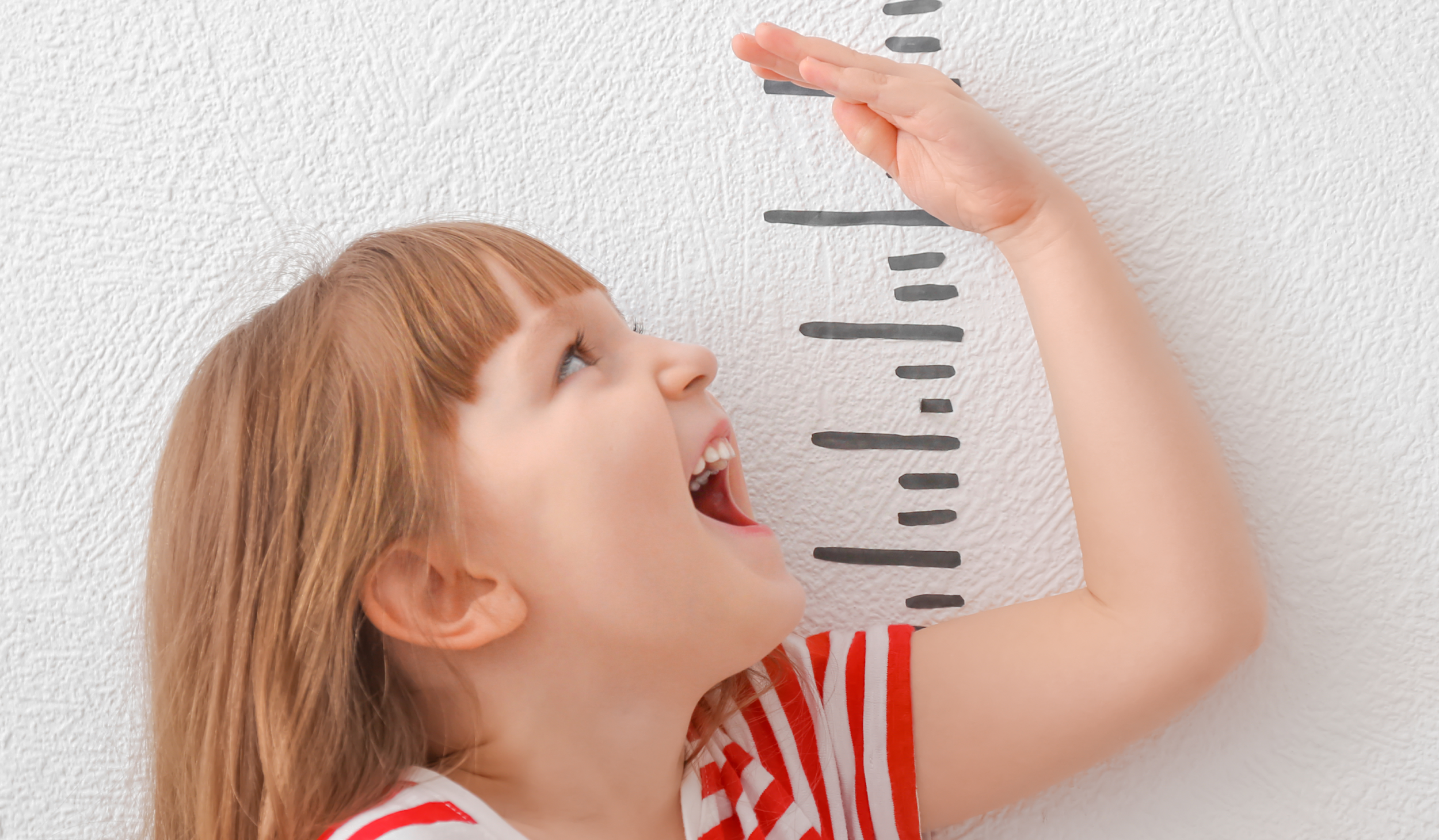 messhoehe-des-netten-maedchens-auf-hellem-hintergrund 2 Ein kleines Mädchen hält ihre Hand an ein selbtgemaltes Maßband, um ihre Größe zu messen. Dabei lacht sie glücklich