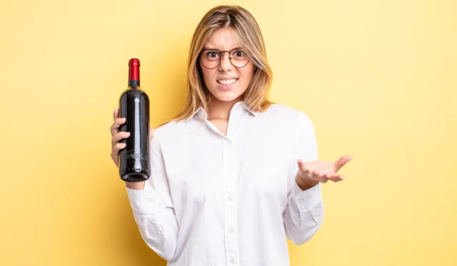 Ein blonde Frau mit Brille und weißem Hemd, die eine Flasche Rotwein hält. Sie schaut den Beobachter skeptisch an.