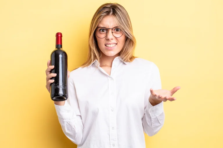 Ein blonde Frau mit Brille und weißem Hemd, die eine Flasche Rotwein hält. Sie schaut den Beobachter skeptisch an.