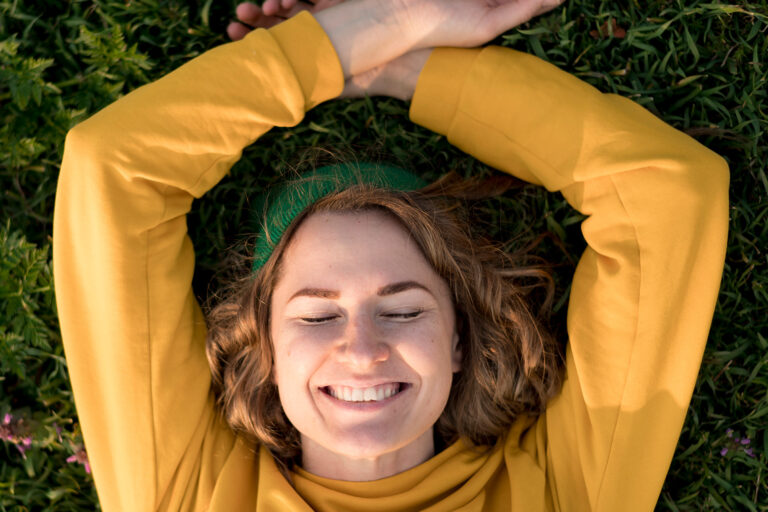 Eine junge Frau mit grünem Haarband und gelbem Pullover liegt entspannt auf einer Wiese und lächelt glücklich